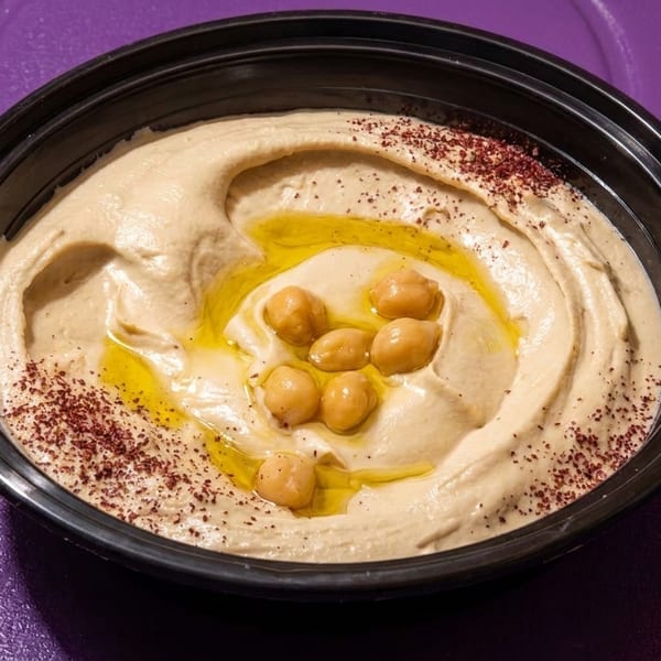 Creamy hummus in a black bowl, drizzled with olive oil, chickpeas, and sumac on a purple surface.