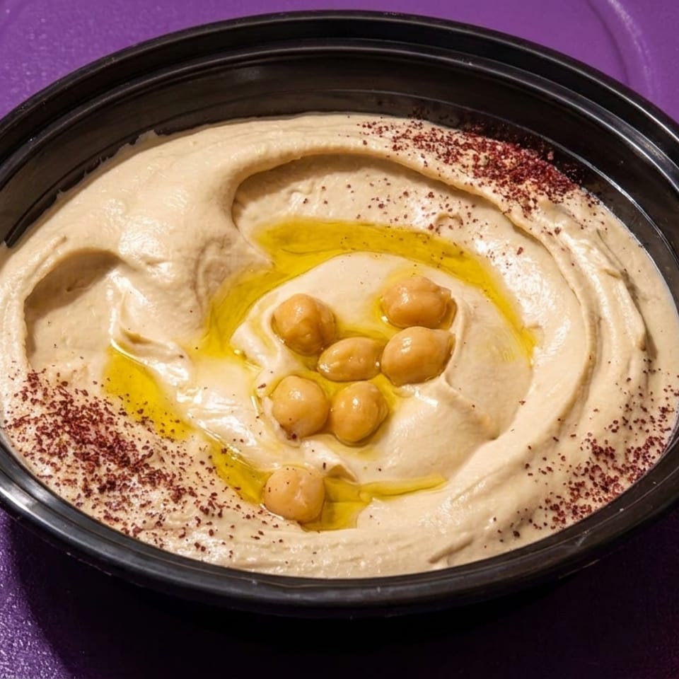 Creamy hummus in a black bowl, drizzled with olive oil, chickpeas, and sumac on a purple surface.