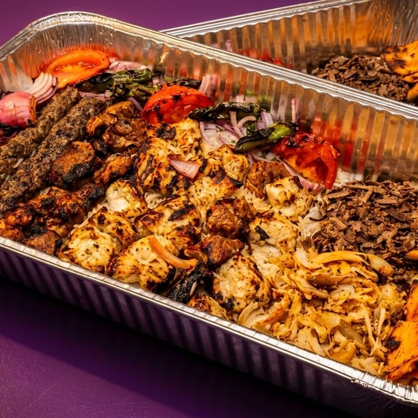 Two large foil trays filled with various grilled meats, rice, vegetables, and pita bread.