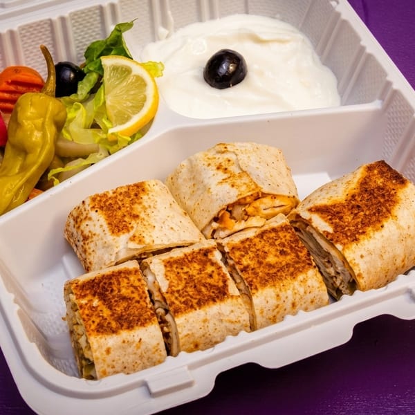 Shawarma wraps, pickles, salad, and white sauce in a white takeout container on a purple surface.